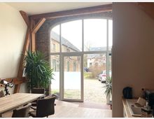 Wohnraum mit großen, bodentiefen Fenstern und einem halbkreisförmigen Oberlicht, durch die ein gepflasterter Hof mit historischen Backsteingebäuden sichtbar ist. Innen befinden sich Holzbalken, ein Esstisch mit Stühlen, Zimmerpflanzen und eine Küchenzeile.