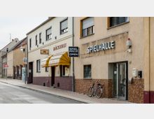 Straßenszene mit einer Bäckerei und einer Spielhalle in einem niedrigen Gebäude mit Fassaden aus hellem Putz und Backstein. Vor der Spielhalle steht ein Fahrrad auf dem Gehweg, und die Bäckerei hat ein gelbes Markisendach.