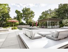 Moderne Terrasse mit großzügigem Poolbereich, umgeben von Bäumen und Sträuchern. Helle Sitzmöbel, ein roter Sonnenschirm, sowie ein minimalistisches Gebäude mit großen Glasfronten bilden die Hauptbestandteile der Anlage.
