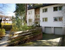 Wohngebäude mit weißer Fassade und mehreren Fenstern, umgeben von Bäumen und Sträuchern. Im Vordergrund befindet sich eine kleine Einfahrt mit zwei Garagentoren und ein Holzzaun, daneben ein gepflasterter Weg.