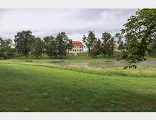 Große Grünfläche mit einem Teich im Vordergrund und einem herrschaftlichen Gebäude mit weißer Fassade und rotem Dach im Hintergrund, umgeben von Bäumen. Der Himmel ist bewölkt, und die Umgebung wirkt wie ein Park oder eine Schlossanlage.