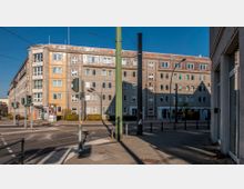 Wohngebäude aus mehreren Stockwerken an einer Straßenecke mit grauer und gelber Fassade. Im Vordergrund befinden sich Straßenbahngleise, Ampeln, Gehwege und Straßenschilder.