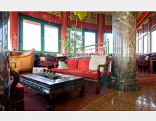 Ein traditionell gestalteter Innenraum mit chinesischem Stil, geprägt von roten und grünen Farbtönen, verzierten Holzmöbeln mit roten Polstern, einem marmornen Pfeiler und großen Fenstern. Im Vordergrund steht ein reich verzierter Holztisch mit Dekoration.