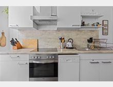 Moderne Küche mit weißen Schränken und grauer Arbeitsplatte, ausgestattet mit einem Backofen, einem Herd, einem Wasserkocher und einer Spüle. An der Rückwand befinden sich ein hölzernes Schneidebrett, ein Messerblock und Küchengeräte in einem Behälter; rechts steht ein Geschirrabtropfgestell.