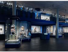 Ein modernes Museum mit beleuchteten Glasvitrinen, die traditionelle Samurai-Rüstungen und Waffen ausstellen. Dunkle Böden, blaue Wände und eine obere Ebene mit weiteren Exponaten verleihen dem Raum eine strukturierte und thematisch konzentrierte Atmosphäre.