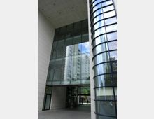 Moderne Gebäudeansicht mit einer großen Glasfassade, die den Blick auf höhere Wohngebäude im Hintergrund reflektiert. Ein überdachter Durchgang führt durch den Eingangsbereich zwischen rechteckigen Steinwänden und einer abgerundeten, verglasten Säule.