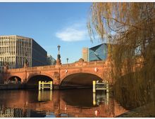 Rote Steinbrücke mit drei Bögen und Laternen, flankiert von modernen Bürogebäuden im Hintergrund; im Vordergrund hängen Äste eines Baumes über einem Fluss. Ein Glaspavillon mit dem DB-Logo ist rechts sichtbar.