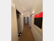 Ein schmaler, gut beleuchteter Flur mit weißen Wänden und einem beigefarbenen Teppichboden. Die Wände sind mit modernen Wandleuchten und einem großformatigen Wandbild mit roten und schwarzen Akzenten dekoriert; mehrere Türen führen zu angrenzenden Räumen.