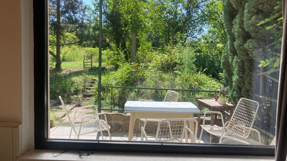 Blick durch ein großes Fenster auf eine Terrasse mit weißem Tisch und mehreren Metallstühlen. Dahinter liegt ein üppig bewachsener Garten mit Bäumen, Sträuchern und einem schmalen Weg.