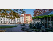 Straßenszene mit einem modernen Gewächshaus auf der rechten Seite, beschriftet mit „Grabpflege und Gestaltung“, umgeben von Pflanzen und Blumenarrangements. Im Hintergrund sind mehrstöckige, farbige Wohnhäuser mit roten Dächern und eine Bushaltestelle zu sehen, eingerahmt von Herbstbäumen.