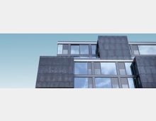 Moderne Fassade eines Bürogebäudes mit großen Glasfenstern und dunklen, rechteckigen Metallverkleidungen unter klarem, blauem Himmel. Das Design zeigt gestaffelte Ebenen mit sichtbaren Reflexionen auf den Fensterscheiben.