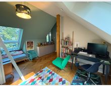 Dachzimmer mit schrägen Wänden, eingerichtet als Kombination aus Schlaf- und Arbeitsbereich. Links ein Bett unter einem Dachfenster, rechts ein Schreibtisch mit Computer und Regalen voller Bücher; Holzboden und ein bunter Teppich ergänzen den Raum.