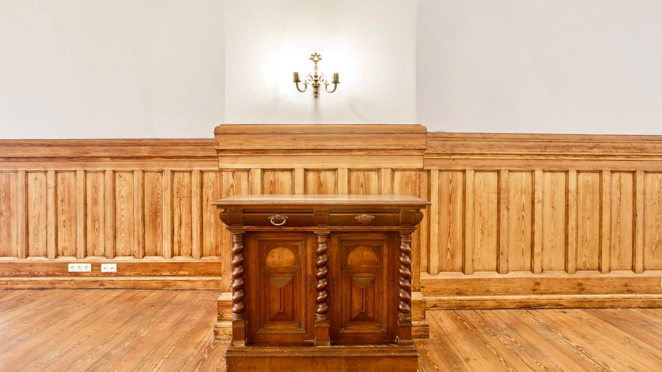 Holzvertäfelter Innenraum mit breiten Dielenboden und weißen Wänden. In der Mitte steht ein dunkles Holzpult mit gedrechselten Säulen, darüber eine Wandlampe im klassischen Stil.