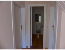 Flur mit drei Türen, von denen die mittlere geöffnet ist und in ein kleines Badezimmer mit Toilette, kleinem Waschbecken und einem Bild an der Wand führt. Der Boden besteht aus Holz, die Wände sind hell gestrichen.