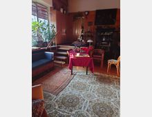 Wohnzimmer mit Holzwänden und gemütlichem Ambiente, ausgestattet mit einem orientalischen Teppich, einer roten Tischdecke auf einem Esstisch, Holzmöbeln und einer Sitzbank. Im Hintergrund befinden sich Pflanzen, Schränke, Regale und dekorative Elemente wie Kerzenhalter und Wandgemälde.