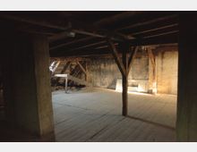 Ein leerer Dachboden mit Holzboden, sichtbaren Holzbalken und schrägen Dachstützen. Die Wände sind größtenteils unverputzt, und durch ein kleines Fenster fällt etwas Licht in den Raum.