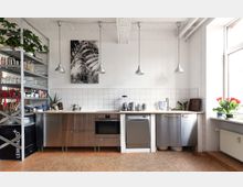 Moderne Wohnküche mit Arbeitsplatte aus Holz, Edelstahlgeräten und einer Rückwand aus weißen Kacheln. Links ein offenes Metallregal mit Geschirr und Pflanzen, rechts ein großes Fenster mit Blumendekoration.