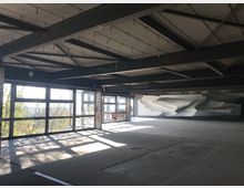 Leerer, unfertiger Innenraum eines Gebäudes mit sichtbaren Stahlträgern und großer Glasfassade, die Tageslicht hereinlässt; Wände sind mit Graffitis und Betontexturen versehen. Im Hintergrund Bäume und Außenblick.