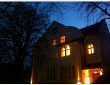 Ein zweistöckiges Gebäude im traditionellen Baustil mit beleuchteten Fenstern und hellem Eingangsbereich, aufgenommen in der Dämmerung. Im Vordergrund ragen dunkle, kahlen Baumäste in den Nachthimmel.