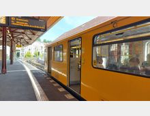 Eine gelbe U-Bahn steht an einem überdachten Bahnsteig mit Holzdecke, daneben ein digitales Anzeigeschild mit Informationen zu den Linien U3 und U1. Im Hintergrund sind Wohngebäude und Bäume sichtbar, während Fahrgäste durch die Fenster des Zuges zu sehen sind.