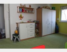 Kinderzimmer mit grüner Teppichauslage, weiß-gelben Wänden und einem Fenster. An der Wand stehen weiße Schränke mit Holzdetails, ein offenes Regal mit Büchern und Spielzeug sowie ein kleines Waschbecken.