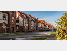 Straßenszene mit einer Reihe historischer Wohnhäuser aus rotem Backstein und weißen Fassaden, ergänzt durch rote Ziegeldächer und dekorative Giebel. Die Straße ist asphaltiert, umgeben von Bürgersteigen, Straßenlaternen und einigen Bäumen.