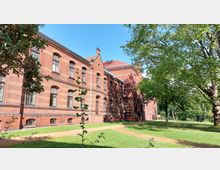 Backsteinbau aus der Gründerzeit mit hohen Fenstern und roten Zierelementen, umgeben von einer Grünanlage mit Rasenflächen, Bäumen und einem sandigen Weg. Der Himmel ist klar und blau.