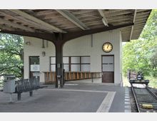 Bahnhofsplattform mit einer überdachten Wartehalle aus hellen Backsteinen, einer Wanduhr, Sitzbänken und einem angrenzenden Gleis. Im Hintergrund sind Bäume und grüne Vegetation zu sehen.