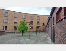 Ziegelbau-Innenhof mit gepflastertem Boden, einem einzelnen Baum, modernen Laternen und Gebäuden mit roten Fensterrahmen. Rechts im Bild befindet sich ein niedrigeres Gebäude mit hellroten Ziegeln.