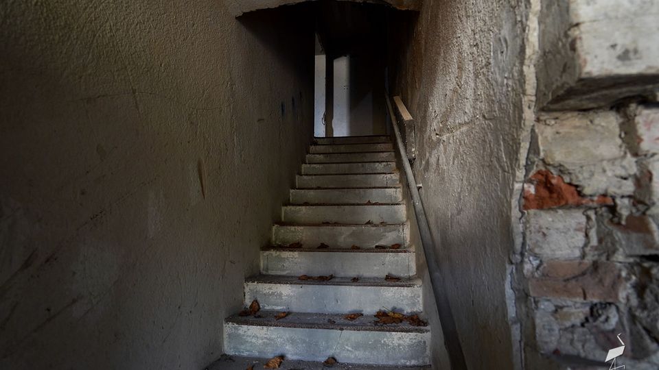 Schmales, dunkles Treppenhaus mit Betonstufen, an den Wänden abgenutzter Putz und rechts ein einfaches Geländer. Oben führt die Treppe durch eine niedrige Öffnung in einen weiteren, schwach beleuchteten Bereich; auf den Stufen liegen trockene Blätter.