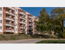 Mehrstöckiges Wohnhaus mit Balkonen, die mit Blumen dekoriert sind, in einer Wohngegend mit Gehweg und Grünflächen im Vordergrund. Im Hintergrund stehen Bäume, die einen parkähnlichen Eindruck vermitteln.