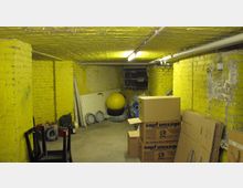 Ein Kellerraum mit gelb gestrichenen Backsteinwänden, einer Betonbodenfläche und sichtbaren Rohrleitungen an der Decke. Der Raum enthält gestapelte Umzugskartons, eine gelbe Gymnastikball, Möbel und verschiedene gelagerte Gegenstände.