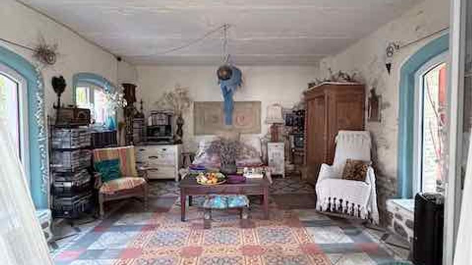 Großer Wohnraum mit bemustertem Fliesenboden und Teppichläufer, weißer Decke und hellen Wänden mit sichtbaren Gebrauchsspuren. In der Mitte stehen ein Couchtisch und Sitzmöbel; entlang der Wände befinden sich Schränke, Kommoden und Dekoration, flankiert von zwei blauen Fensterrahmen.