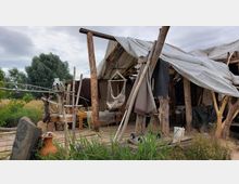 Eine einfache, rustikale Hütte aus Holz und Planen, umgeben von hohem Gras und Bäumen. Vor der Hütte befinden sich ein hängender Stoffstuhl, Holzmöbel und dekorative Elemente wie ein Tierknochen und Tongefäße.