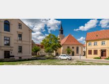 Kleiner Platz in einer dörflichen Umgebung mit einer Kirche im Zentrum, erkennbar an ihrem roten Dach und Glockenturm, flankiert von historischen Wohngebäuden mit gelben und beige Fassaden. Im Vordergrund stehen ein Baum und ein geparktes weißes Auto.