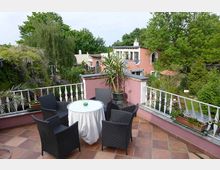 Eine Terrasse mit Terrakottafliesen, einem runden Tisch mit weißem Tuch und vier schwarzen Korbsesseln. Im Hintergrund sind ein grüner Garten mit dichtem Pflanzenbewuchs und mehrere pastellfarbene Gebäude mit großen Fenstern sichtbar.