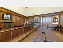 Holzvertäfelter Raum mit Sitzungssaalcharakter, ausgestattet mit einem langen Tisch, Stühlen und Mikrofon. An den Wänden hängen Gemälde und ein Wandteppich mit Wappen; außerdem befinden sich dort Flaggen und Wandlampen.