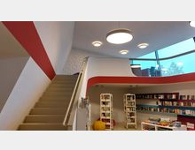 Modernes Bibliotheksinterieur mit einer geschwungenen Treppe, einer roten und weißen Wandgestaltung sowie einer offenen Galerie. Im Erdgeschoss befinden sich Bücherregale, Spiele und dekorative Elemente, während die obere Etage durch eine gläserne Brüstung und große Fenster mit Blick nach außen begrenzt ist.