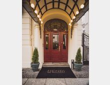 Eingangsbereich eines klassischen Hotels mit zwei roten Holztüren unter einem gewölbten Vordach mit Beleuchtung. Links und rechts des Eingangs stehen grüne Pflanzkübel, und auf der schwarzen Fußmatte ist der Schriftzug "HENRI Hotel Berlin Kurfürstendamm" zu sehen.