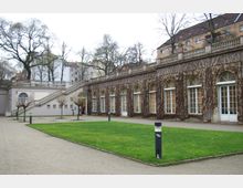 Außenansicht eines historischen Gebäudes mit mehreren bogenförmigen Fenstern, von Kletterpflanzen bewachsen, umgeben von einem kleinen Park mit Grünflächen und Gehwegen. Rechts führt eine breite Treppe zu einer erhöhten Terrasse, während im Hintergrund weitere Stadthäuser sichtbar sind.