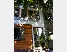 Moderne Fassade eines Wohnhauses mit einer Kombination aus Holz- und Metallverkleidung, großen Fenstern und grünen Fensterläden. Im Vordergrund sind Schatten von Bäumen sichtbar, die auf die Hauswand fallen.