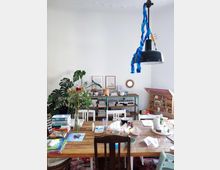 A casual dining or workspace with a wooden table surrounded by mismatched chairs, cluttered with books, papers, and craft items. The background features a white wall, a green shelving unit filled with books and decor, a large leafy plant, and a hanging black lamp with blue streamers.