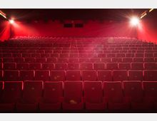 Innenraum eines Kinosaals mit mehreren Reihen roter, nummerierter Kinositze, gleichmäßig ausgerichtet. Im Hintergrund sind zwei beleuchtete Lampen an den Wänden zu sehen, die den Raum leicht erhellen.