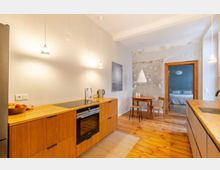 Offene Küche mit minimalistischer Einrichtung, gekennzeichnet durch Holzfronten, eine Arbeitsplatte mit Kochfeld und eingebautem Backofen. Im Hintergrund ein kleiner Essbereich mit rundem Holztisch und Stühlen vor einer Wand mit unvollständig verputzter Optik; durch eine offene Tür ist ein Schlafzimmer sichtbar.