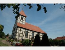 Fachwerkkirche mit rotem Satteldach und Glockenturm, umgeben von einem gepflegten Friedhof mit Blumen und kleinen Bäumen, unter einem klaren blauen Himmel.