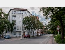 Straßenszene mit einem vierstöckigen Wohngebäude aus grauem Putz an einer Kreuzung, umgeben von Bäumen. Auf der Straße stehen geparkte Autos, und rechts ist ein Geh- und Radweg sichtbar, daneben ein Warnschild für Straßenschäden.