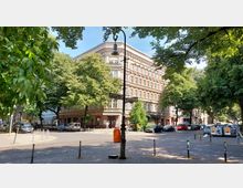 Straßenkreuzung in einer Stadt mit einem historischen mehrstöckigen Wohngebäude im Hintergrund, umgeben von dichtem Grün großer Straßenbäume. Im Vordergrund stehen Straßenschilder und ein orangener Mülleimer, während Autos entlang des gepflasterten Straßenrands parken.