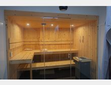 Eine Sauna aus hellem Holz mit zwei Sitzebenen und einer gläsernen Eingangstür. Im Vordergrund steht ein elektrischer Saunaofen, und die dunklen Bodenfliesen bilden einen Kontrast zur Holzvertäfelung.