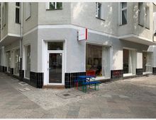 Eckgebäude mit heller Fassade und schwarzen Fliesen am unteren Bereich, großen Schaufenstern und einer Glastür. Vor dem Gebäude stehen blaue und grüne Kindersitzmöbel auf einem gepflasterten Gehweg in einer städtischen Umgebung.