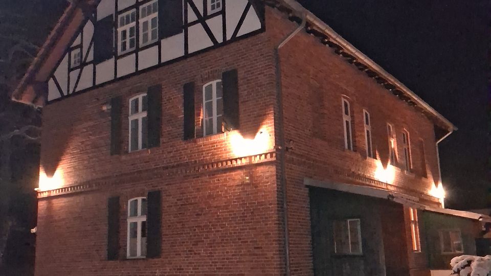 Backsteingebäude im Fachwerkstil mit beleuchteten Fenstern und weißem Schnee im Vordergrund, aufgenommen bei Nacht. Das obere Geschoss zeigt ein Fachwerkdesign, während die unteren Fassaden aus unverputztem Ziegelstein bestehen.